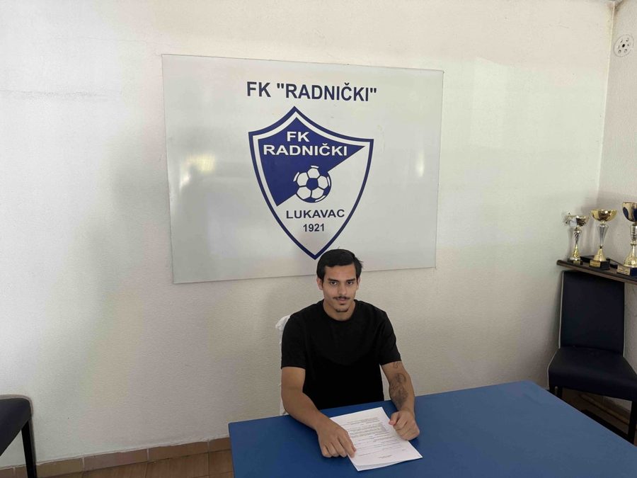 Mohamed Almosawi signing paperwork beneath the FK Radnicki Lukavac crest in Bosnia and Herzegovina